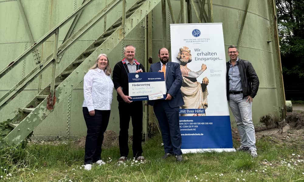Vertreter der Deutschen Stiftung Denkmalschutz und der Stadtwerke Dinslaken stehen neben einem Roll-up vor dem Gasometer der Stadtwerke