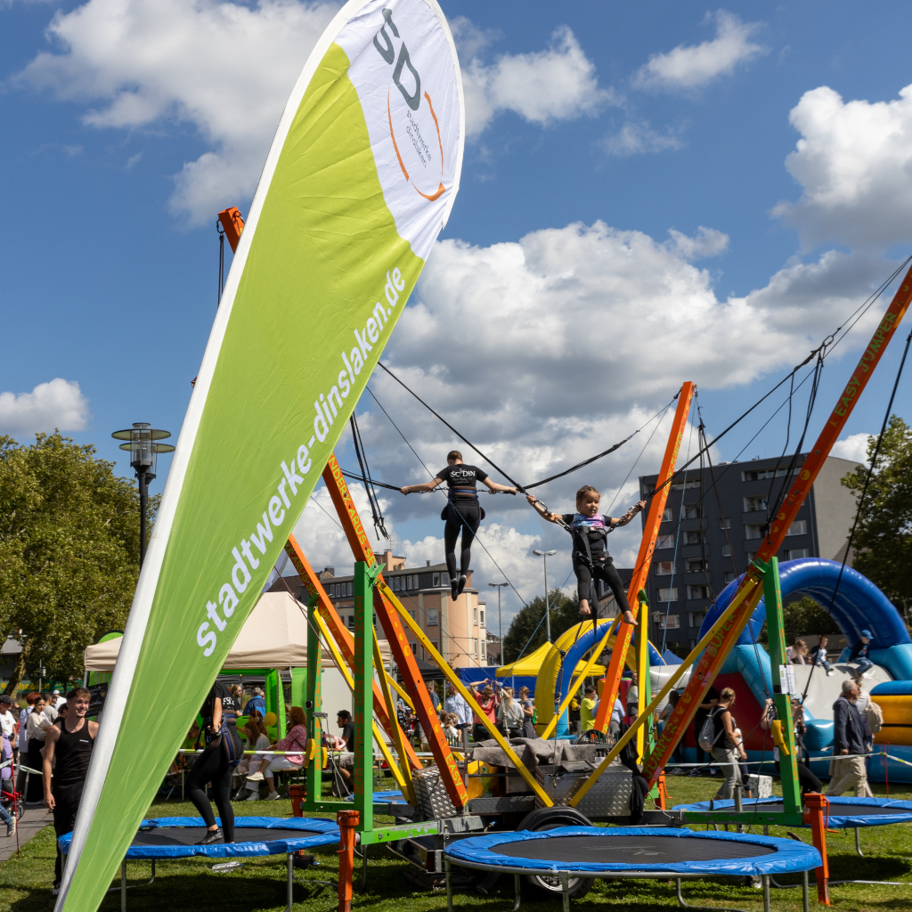 Kinder springen auf dem Bungee-Trampolin bei den Jugend-DIN-Tagen im Stadtpark. Im Vordergrund ragt ein Beach-Flag der Stadtwerke Dinslaken ins Bild