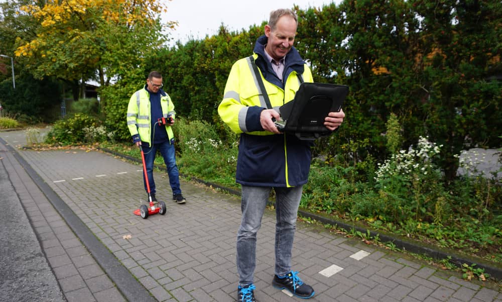 Zwei Stadtwerke-Mitarbeiter gehen einen Gehweg mit Sonde und Laptop ab.