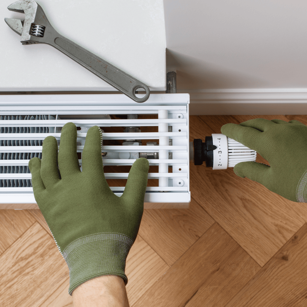 Eine Hand und ein Schraubenschlüssen liegen oben auf der Heizung, die andere Hand ist am Heizkörperventil.