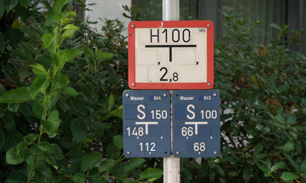 Die Beschilderung eines Unterflurhydranten an einer Stele vor hohen Sträuchern.