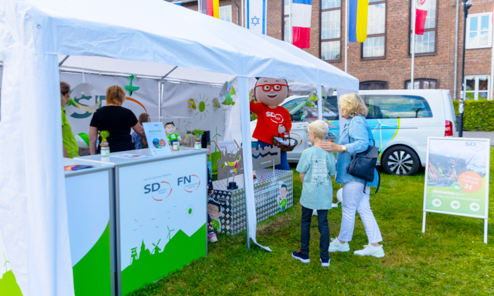 Das Maskottchen DIN begrüßt ein Kind in Begleitung einer Erwachsenen am Stand der Stadtwerke Dinslaken