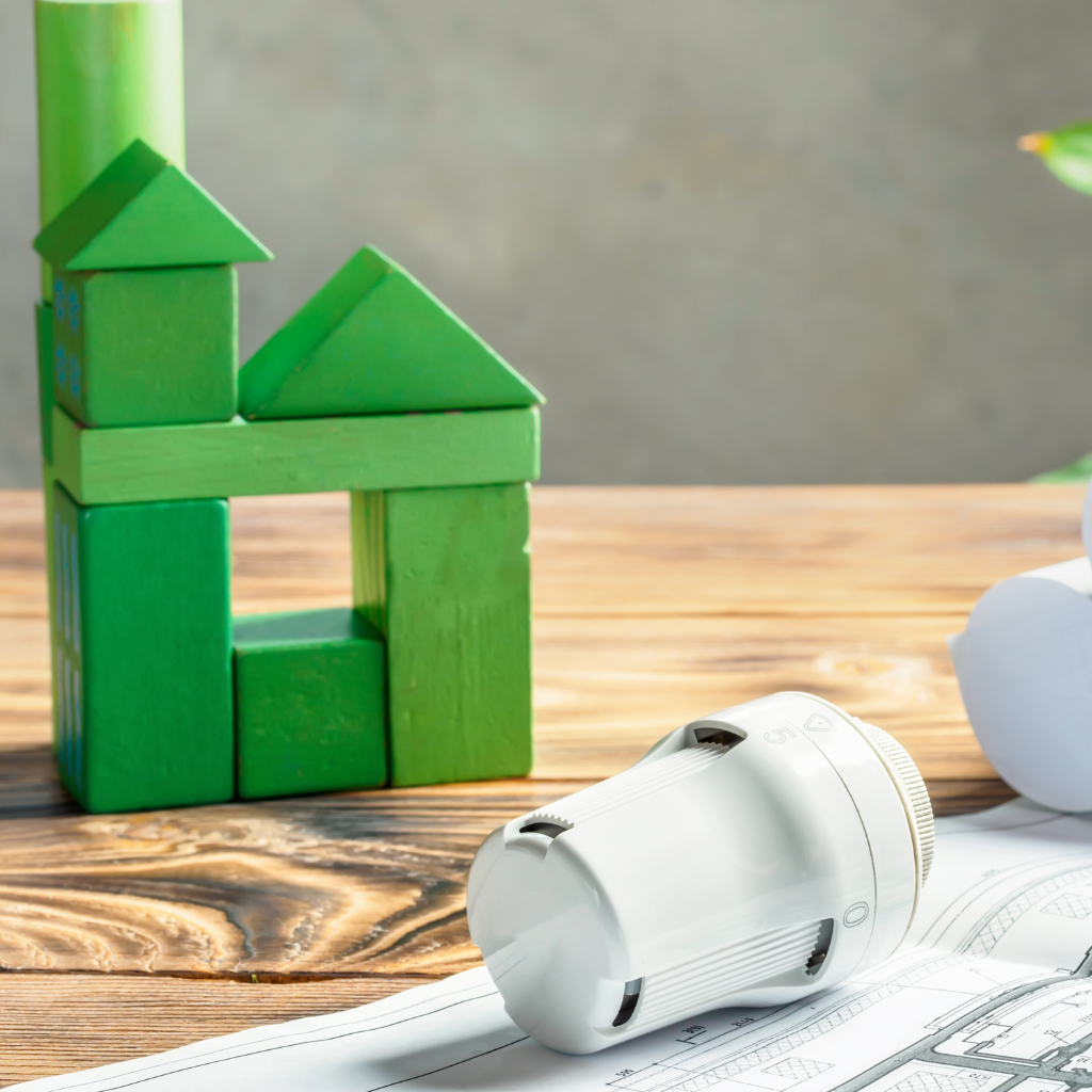 Thermostatisches Heizkörperventil und grünes Würfelhaus auf Holzuntergrund mit Bauzeichnungen